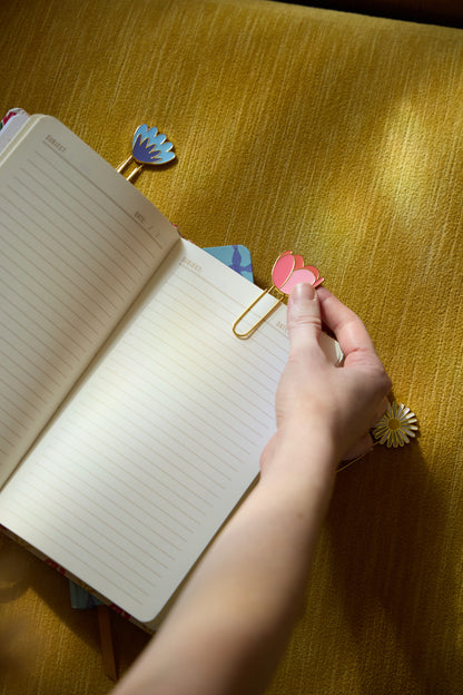 A hand attaches a pink floral paperclip from the Set of 3 Paperclips - Floral to an open lined notebook on a yellow textured surface, with other colorful clips from the set scattered nearby.