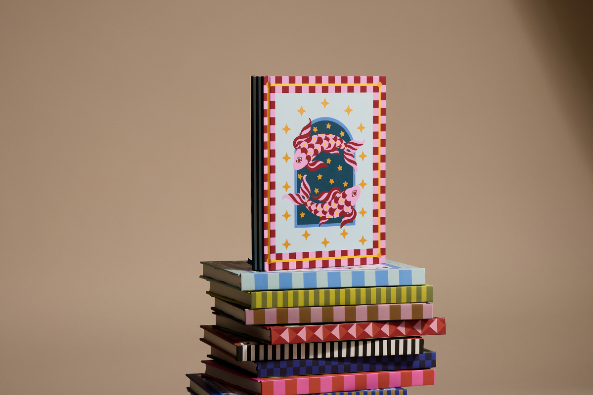 A stack of colorful patterned books with a square art book standing upright on top. The top book features a red-and-white check border and an illustration of two pink and blue fish on a light background.