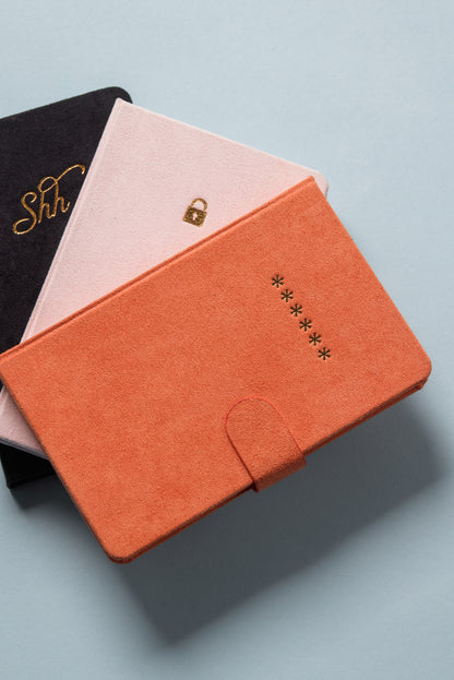 Three suede-covered notebooks are stacked on a blue surface. On top is the Mini Password Journal - Asterisks in orange with gold asterisks, followed by a pink notebook with a gold lock, and a black one labeled “Shh” in gold script.