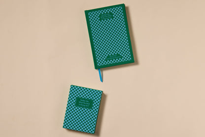 Two green and blue checkered Pardon Me Journals with dark green borders feature bookcloth covers, lay-flat binding, and a blue ribbon bookmark. Both display text in a small green rectangle on the cover, shown on a beige surface.