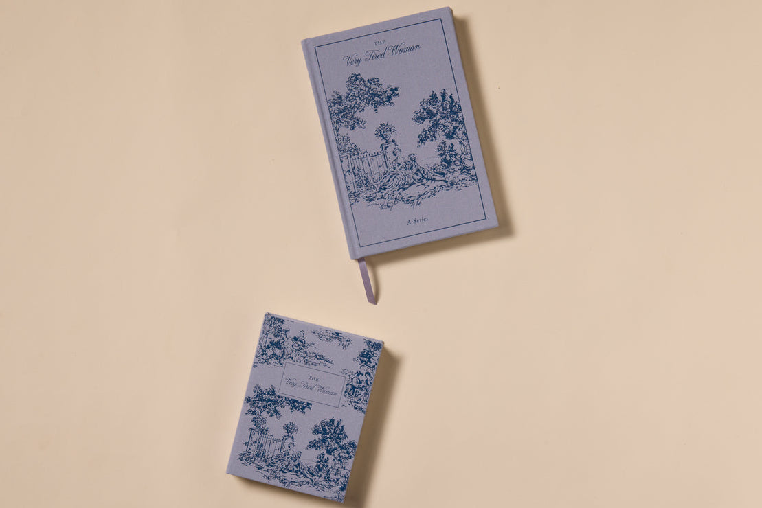 Two "The Very Tired Woman Journal" notebooks with blue vintage illustrations rest on a beige background. The larger, lay-flat edition with a ribbon bookmark sits above the smaller, both evoking vintage book journal charm.
