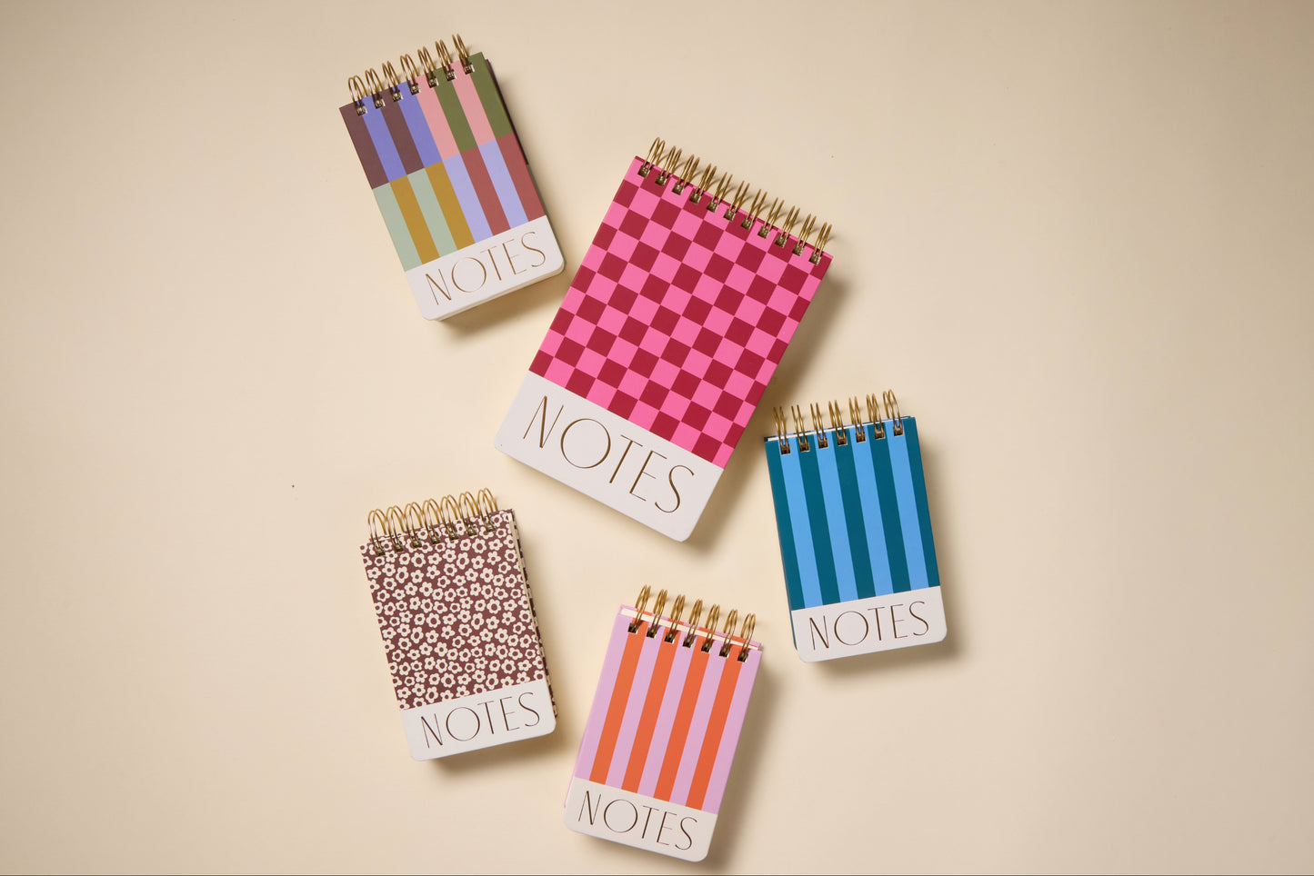 Five Chunky Large Twin Wire Notepads in Burgundy and Pink, each spiral-bound and labeled NOTES, with patterns like stripes, checks, and circles. Made with acid-free paper and soy-based ink, they're arranged neatly on a beige surface.