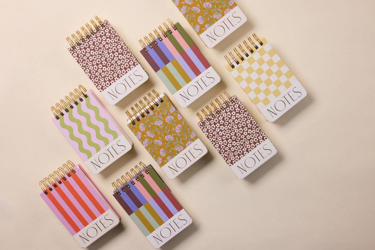 Nine Chunky Small Twin Wire Notepads - Checks, each with a colorful checkered cover and "NOTES" at the bottom, are neatly arranged on a beige background.