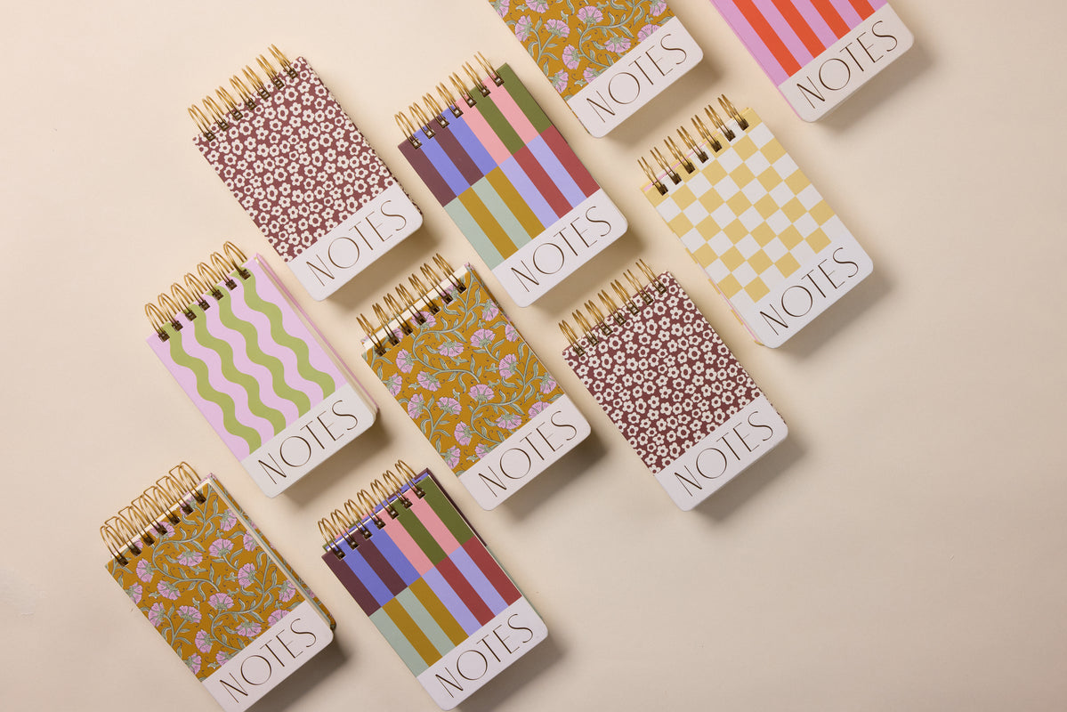 Nine Chunky Small Twin Wire Notepads – Wavy Stripes, each with NOTES on the cover, are arranged on a beige surface. The notepads display unique, colorful designs with floral, checkered, striped, wavy stripes, and circular patterns.