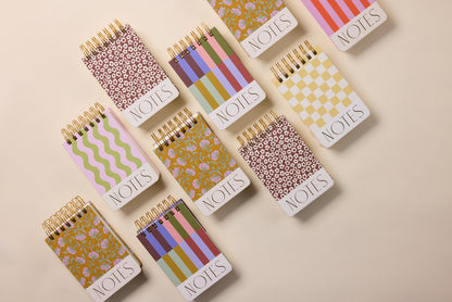 Nine Chunky Small Twin Wire Notepads – Wavy Stripes, each with NOTES on the cover, are arranged on a beige surface. The notepads display unique, colorful designs with floral, checkered, striped, wavy stripes, and circular patterns.