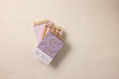 A Large Chunky Notepad - Purple Vintage Floral with a hard cover and "NOTES" written on the front rests atop two other spiral-bound, lined notepads with pastel and gold floral designs, all stacked on a beige surface.