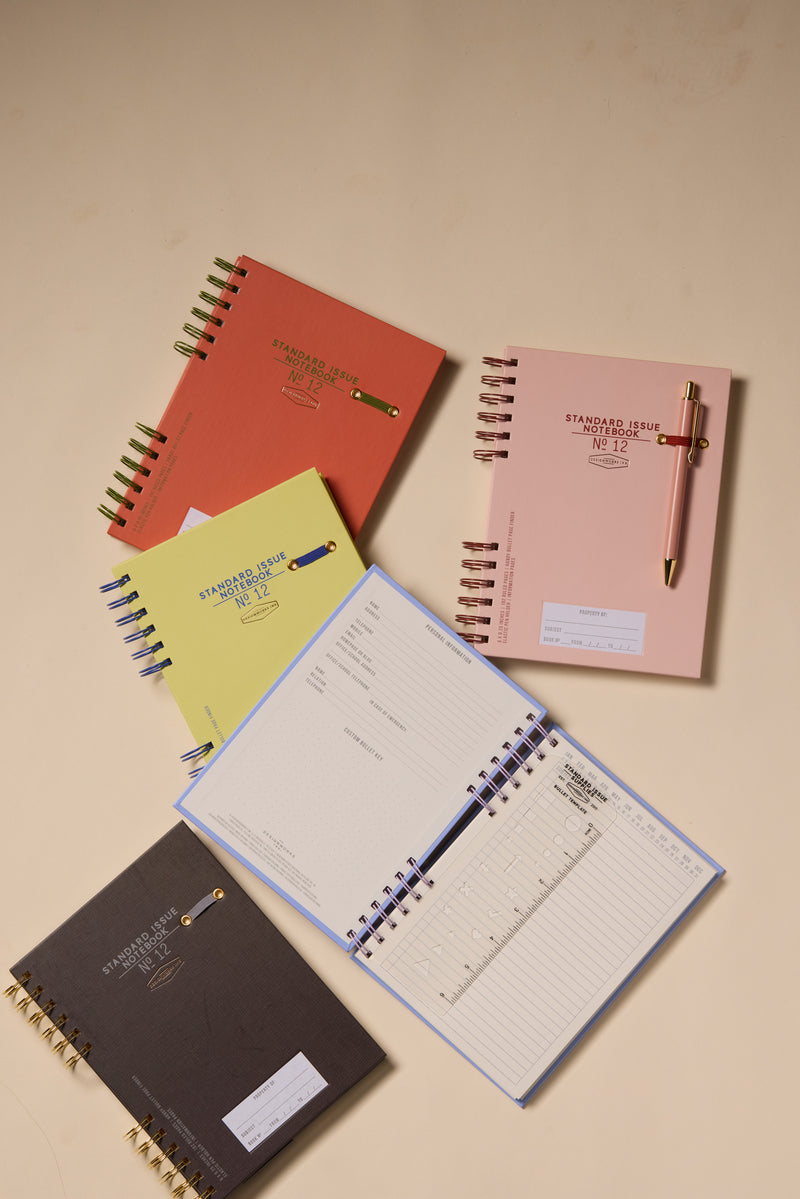 The Standard Issue Notebook No.12 in Dusty Pink is displayed on a beige surface, paired with other colorful notebooks. Two planners are open to lined and calendar pages, while a gold pen rests atop the Dusty Pink notebook.