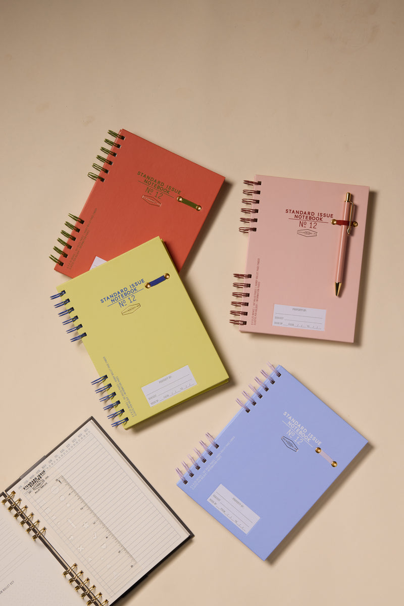The Standard Issue Planner Notebook No. 12 - Rosewood + Blush is displayed open to reveal lined and bullet pages, offering a premium writing experience, alongside other colorful spiral-bound planners with pens clipped on top.