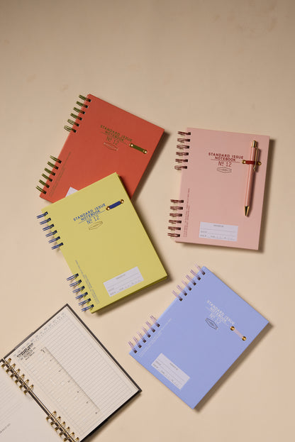 The Standard Issue Planner Notebook No. 12 - Rosewood + Blush is displayed open to reveal lined and bullet pages, offering a premium writing experience, alongside other colorful spiral-bound planners with pens clipped on top.