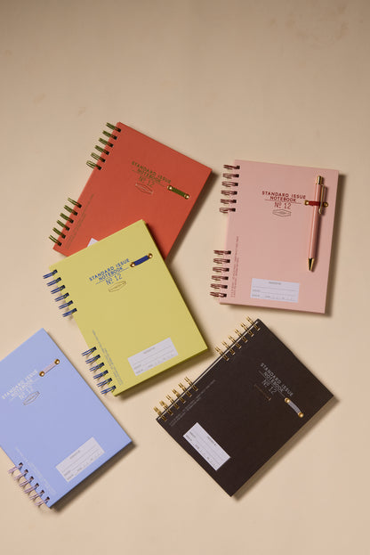 Two Standard Issue Planner Notebook No. 12 in Army Green and Chili are displayed on a beige surface, with pens clipped to their covers for a premium writing experience.