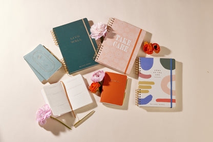 A flat lay of the Guided Wellness Journal - "Come As You Are" with a spiral binding, surrounded by flowers on a light beige background—perfect for your daily gratitude practice.