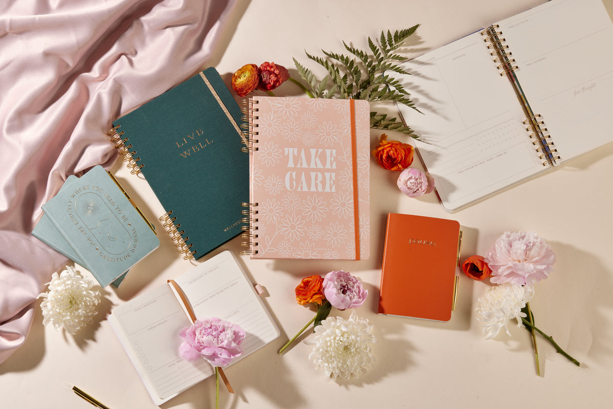 A flat lay of assorted planners, notebooks, and the Guided Wellness Journal - "Take Care," some open and some closed, surrounded by colorful flowers, green fern leaves, and draped pink fabric on a cream surface.