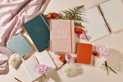 A flat lay of assorted planners, notebooks, and the Guided Wellness Journal - "Take Care," some open and some closed, surrounded by colorful flowers, green fern leaves, and draped pink fabric on a cream surface.