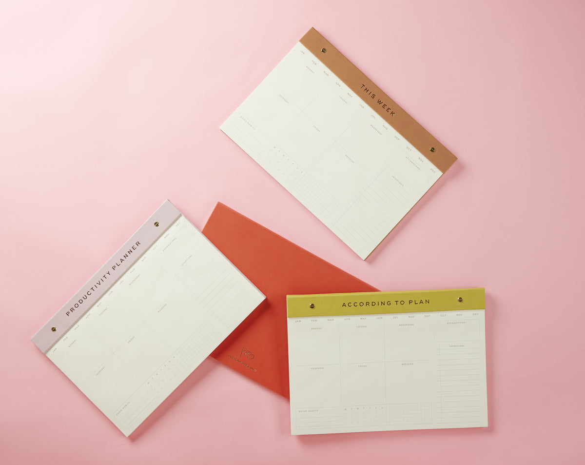 The Weekly Postbound Notepad - Matcha, featuring colored headings for Productivity Planner, This Week, and According to Plan, with a red envelope, is arranged on a pink background—perfect as a stylish desk organizer.