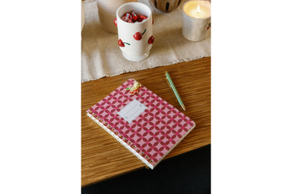 A Tulip Small Twin Wire Notebook in Pinwheel Pink, a green pen, a cherry-patterned mug, and a lit candle are arranged on a wooden table covered with a beige cloth runner.