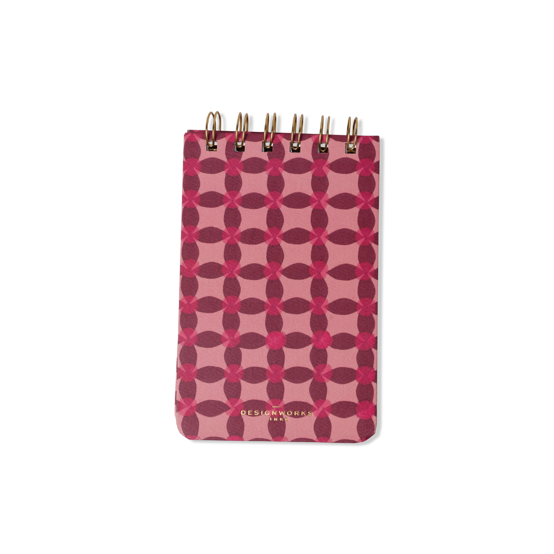 The Tulip Twin Wire Notepad - Pinwheel Pink has a pink and maroon geometric cover, lined pages, gold twin-wire binding at the top, and "DESIGNWORKS" embossed in gold at the bottom.