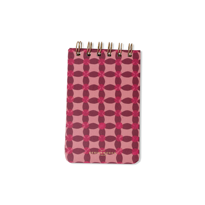 The Tulip Twin Wire Notepad - Pinwheel Pink has a pink and maroon geometric cover, lined pages, gold twin-wire binding at the top, and "DESIGNWORKS" embossed in gold at the bottom.