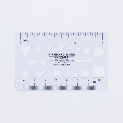 A clear plastic stencil labeled STANDARD ISSUE SUPPLIES BULLET TEMPLATE with geometric shapes and rulers (inches/centimeters), designed for a premium writing experience with the Standard Issue Notebook No.3 - Blue.