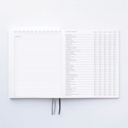 The Standard Issue Notebook No.3 - Black is open to a blank notes page on the left and a multi-year holiday overview on the right, featuring two ribbon bookmarks for a premium writing experience inspired by classic design.